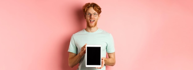 Photo gratuite jeune homme excité aux cheveux rouges et à la barbe vérifiant la promotion en ligne montrant des éboulis de tablette numérique