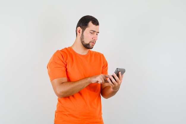 Jeune homme faisant des calculs sur la calculatrice en t-shirt orange et regardant occupé, vue de face.