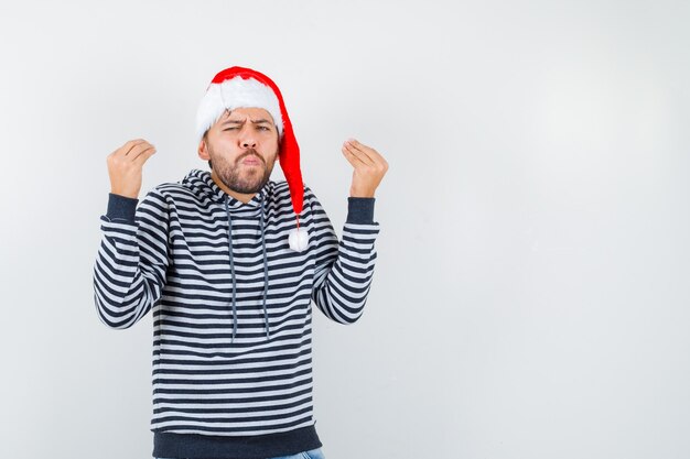 Jeune homme faisant un geste italien, faisant la moue des lèvres en sweat à capuche, bonnet de Noel et ayant l'air confus,