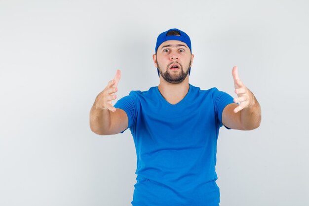 Jeune homme faisant des gestes comme tenant quelque chose de lourd en t-shirt bleu et vue de face de la casquette.