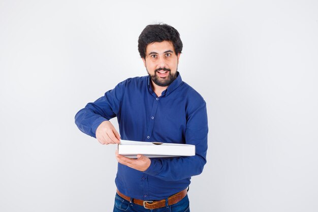 Jeune homme faisant semblant d'ouvrir une boîte à pizza en chemise, jeans et l'air heureux, vue de face.