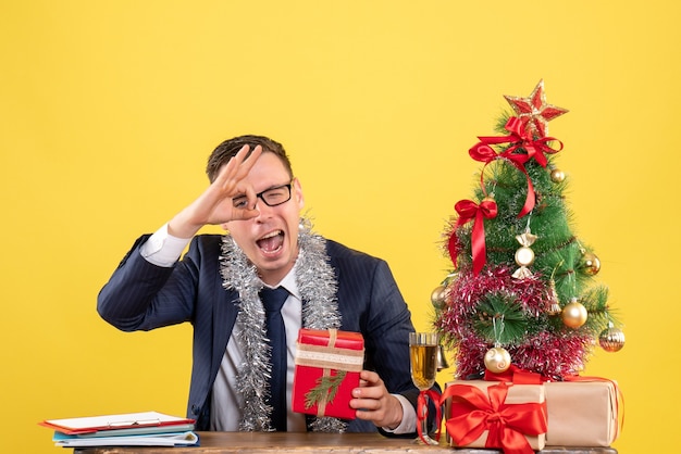 Jeune homme faisant signe ok devant les yeux assis à la table près de l'arbre de Noël et présente sur jaune