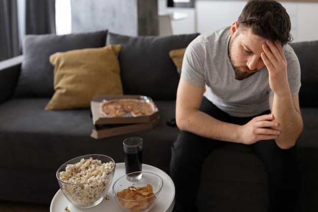 Jeune homme fatigué sur le canapé à la maison assis à côté de la malbouffe