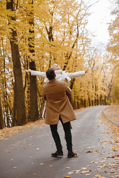 Jeune homme et femme marchant dehors portant des manteaux beiges. Femme blonde et homme brune dans la forêt d'automne. Homme tenant sa femme dans les mains.