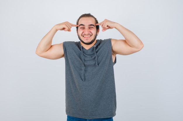 Jeune homme en forme gardant les doigts sur les tempes dans un sweat à capuche sans manches et à la recherche de bonheur. vue de face.