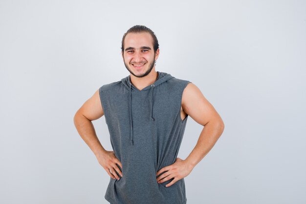 Jeune homme en forme posant avec les mains sur la taille en sweat à capuche sans manches et à la joyeuse vue de face.