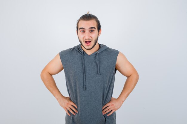 Jeune homme en forme posant avec les mains sur la taille en sweat à capuche sans manches et à la vue de face, choqué.