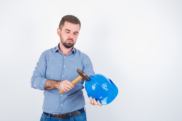 Jeune homme frappant le casque avec un marteau en chemise, jeans, casque et à la grave, vue de face.