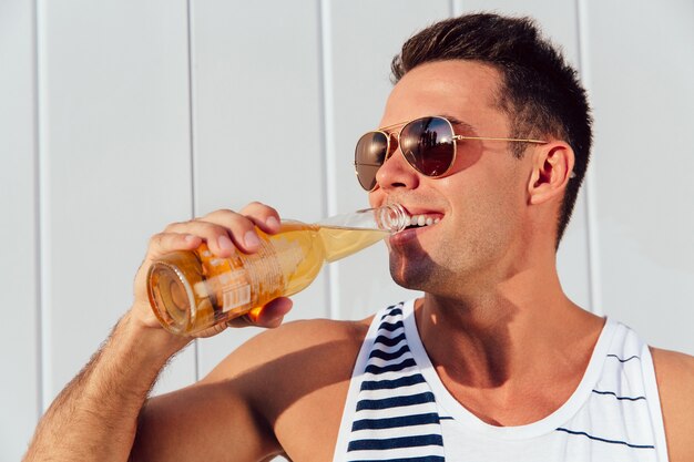 Jeune homme gai dans les lunettes de soleil boire de la bière, tout en se tenant près du mur