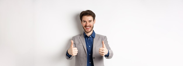 Photo gratuite jeune homme gai et positif en costume d'affaires montrant les pouces vers le haut et souriant comme quelque chose de bien p