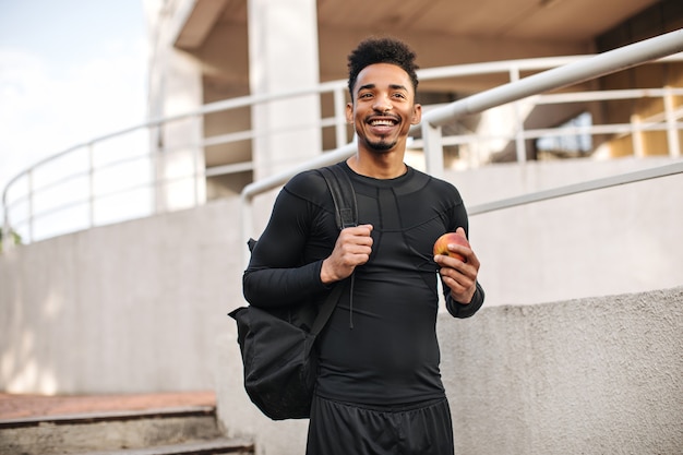 Un jeune homme gai en t-shirt noir à manches longues et en short sourit largement, tient un sac à dos et une pomme, pose à l'extérieur