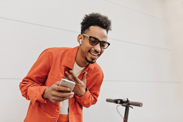 Photo gratuite jeune homme gai en veste orange, lunettes de soleil et t-shirt coloré rit et tient le téléphone