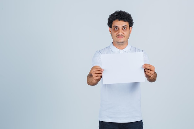 Jeune homme gardant une feuille de papier vierge en t-shirt blanc, pantalon et l'air confiant, vue de face.