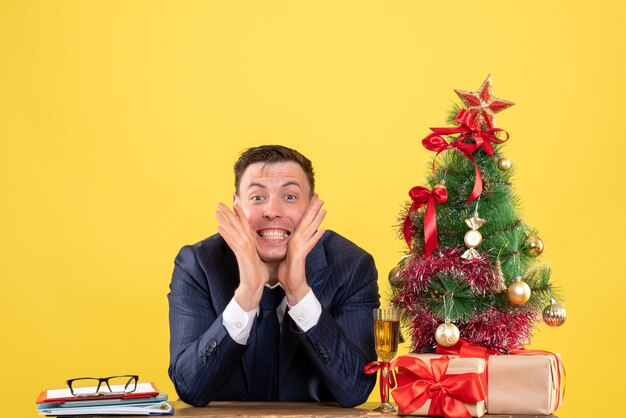Jeune homme gardant des hadns près de la joue assis à la table près de l'arbre de Noël et présente sur jaune