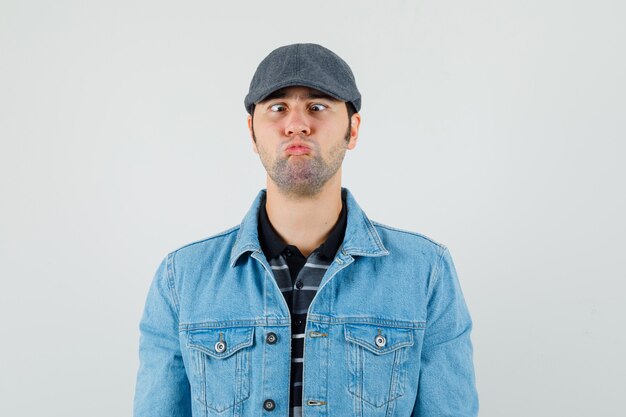 Jeune homme gardant les lèvres pliées avec des yeux strabisme en t-shirt, veste, casquette et à la drôle