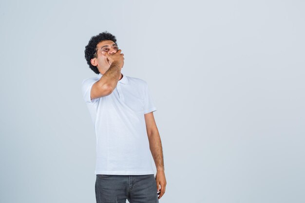 Jeune homme gardant la main sur la bouche en t-shirt blanc, pantalon et l'air heureux. vue de face.