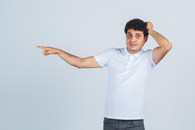 Jeune homme gardant la main sur la tête, pointant de côté en t-shirt blanc, pantalon et l'air perplexe. vue de face.