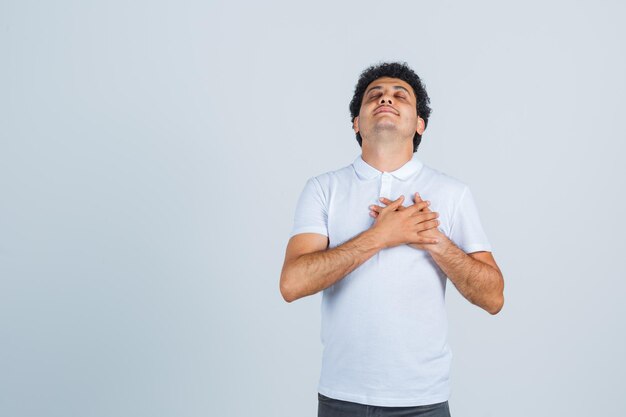 Jeune homme gardant les mains sur la poitrine en t-shirt blanc, pantalon et l'air heureux, vue de face.