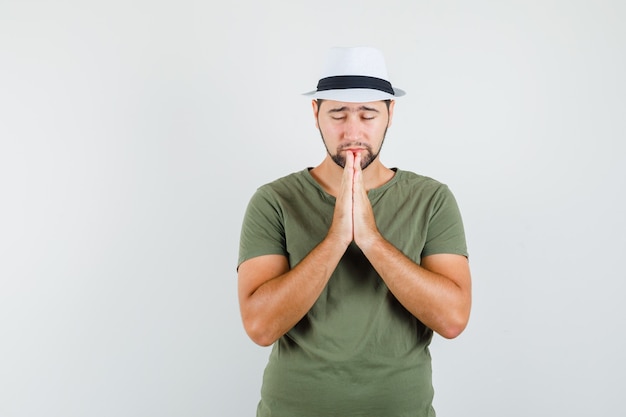 Jeune homme gardant les mains en priant le geste en t-shirt vert et chapeau et à la recherche d'espoir