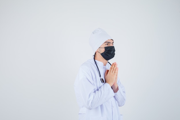 Jeune homme gardant les mains en signe de prière en uniforme blanc, masque et à la recherche d'espoir.