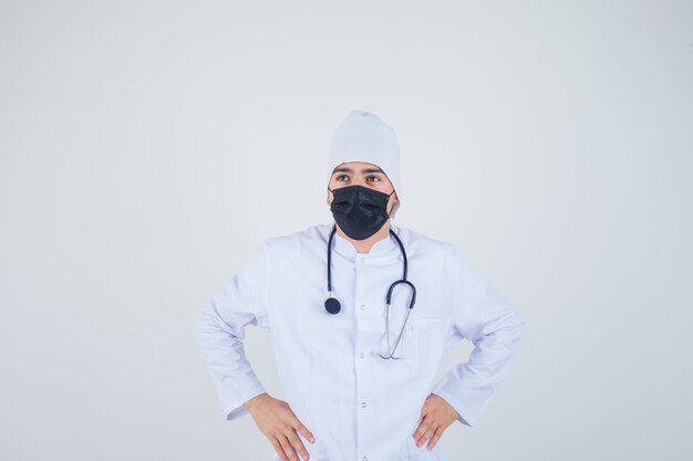 Jeune homme gardant les mains sur la taille en uniforme blanc, masque et à la réflexion.