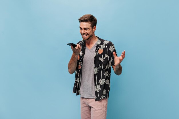 Un jeune homme grincheux en chemise noire à la mode, un t-shirt gris et un short moderne parle avec colère au téléphone sur fond bleu isolé