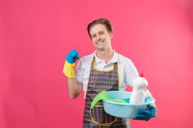 Jeune homme hansdome portant un tablier et des gants en caoutchouc tenant bassin avec des outils de nettoyage souriant pointant vers quelque chose derrière avec le pouce souriant debout sur le mur rose