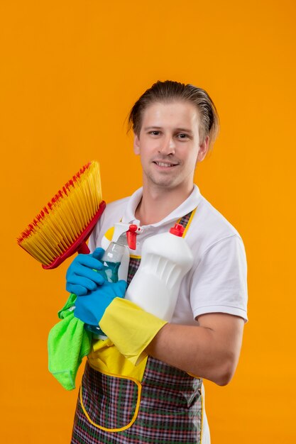 Jeune homme hansdome portant un tablier et des gants en caoutchouc tenant des outils de nettoyage