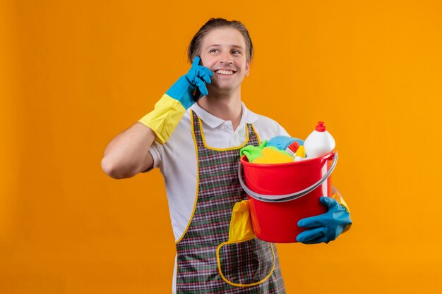 Jeune homme hansdome portant un tablier et des gants en caoutchouc tenant un seau avec des outils de nettoyage souriant joyeusement tout en parlant au téléphone mobile debout sur un mur orange