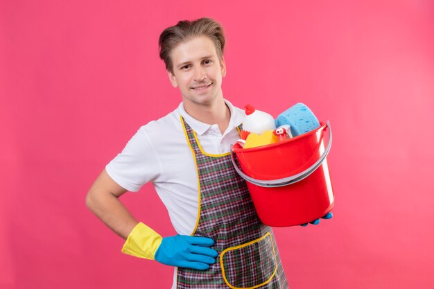 Jeune homme hansdome portant un tablier et des gants en caoutchouc tenant un seau avec des outils de nettoyage avec un sourire confiant sur le visage debout sur un mur rose