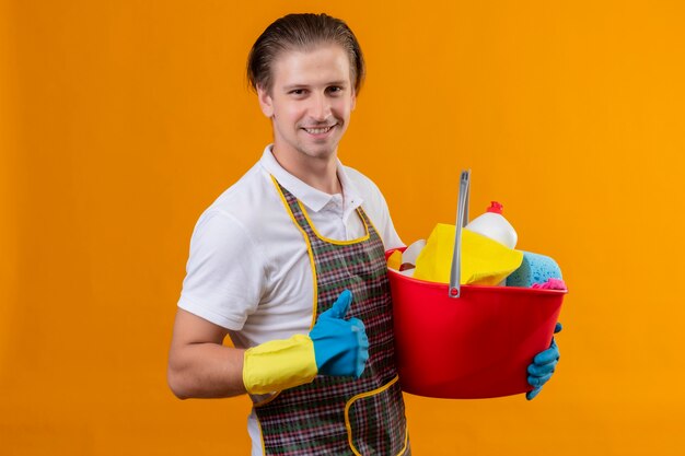 Jeune homme hansdome portant un tablier et des gants en caoutchouc tenant un seau avec des outils de nettoyage