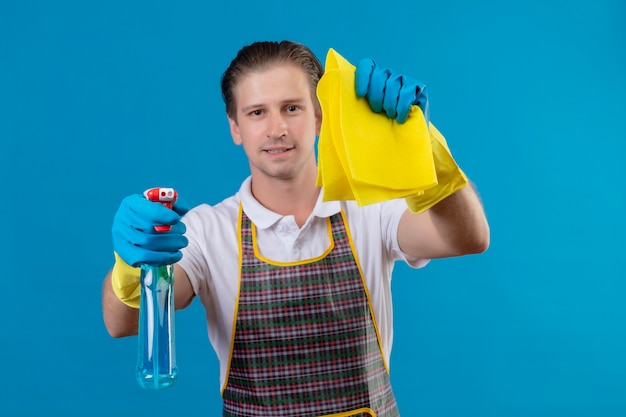 Jeune homme hansdome portant un tablier et des gants en caoutchouc tenant un spray de nettoyage et un tapis