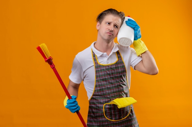 Jeune Homme Hansdome Portant Un Tablier Et Des Gants En Caoutchouc Tenant Une Vadrouille Et Une Bouteille De Produits De Nettoyage