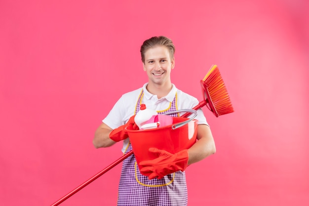 Jeune homme hansdome portant un tablier tenant un seau avec des outils de nettoyage et une vadrouille souriant confiant debout sur un mur rose