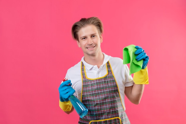 Jeune homme hansdome portant un tablier tenant un spray de nettoyage et un tapis heureux et positif souriant prêt à nettoyer debout sur un mur rose