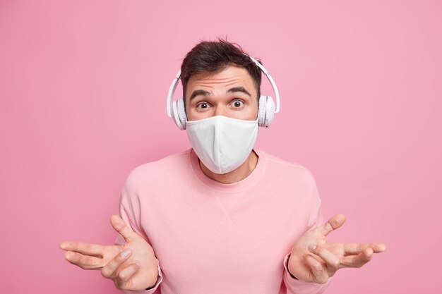 Un jeune homme hésitant sans idée écarte les paumes l'air confus écoute de la musique via des écouteurs sans fil reste à la maison pour ne pas propager la maladie du coronavirus porte un masque de protection