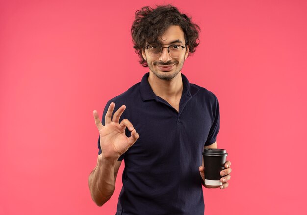 Jeune homme heureux en chemise noire avec des lunettes optiques détient une tasse de café et des gestes ok signe de la main isolé sur un mur rose