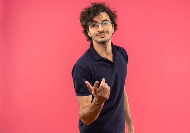 Jeune homme heureux en chemise noire avec des lunettes optiques tient la main isolé sur un mur rose