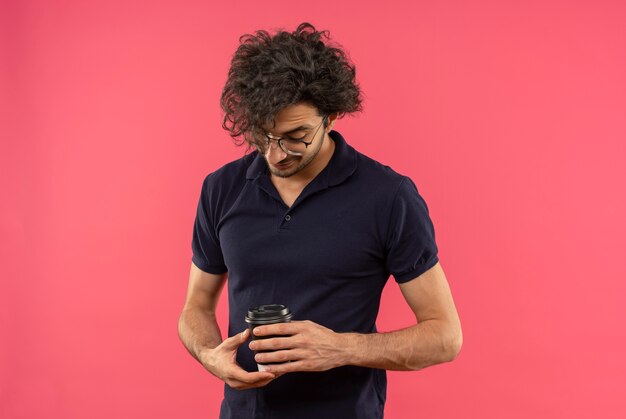 Jeune homme heureux en chemise noire avec des lunettes optiques tient et regarde la tasse de café isolé sur le mur rose