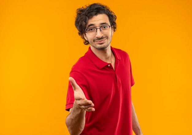 Jeune homme heureux en chemise rouge avec des lunettes optiques tient la main isolé sur un mur orange