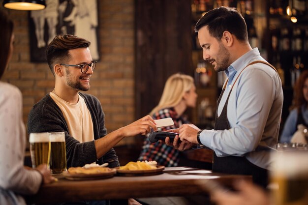 Jeune homme heureux avec un smartphone payant la facture par paiement sans contact dans un bar