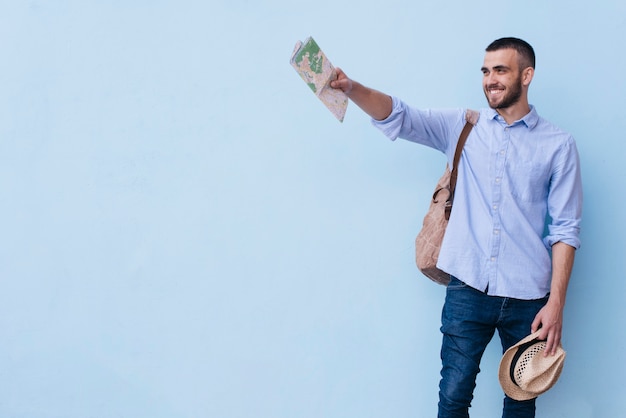 Jeune homme heureux tenant la carte et chapeau montrant quelque chose debout sur fond bleu