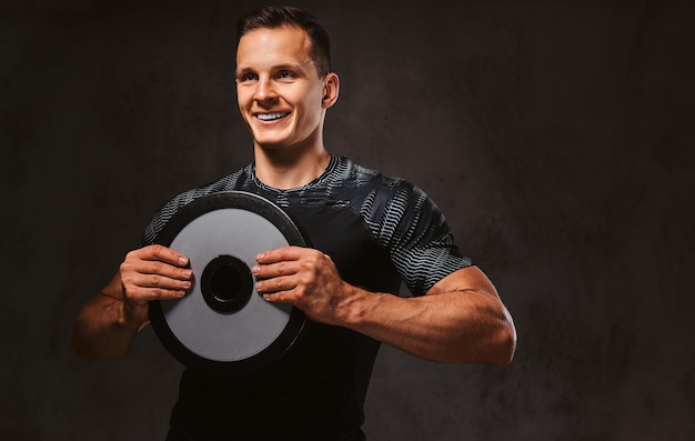 Un jeune homme heureux en tenue de sport tenant un disque d'haltères sur fond sombre.