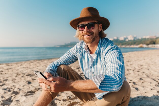 Jeune homme hipster attrayant assis sur la plage au bord de la mer en vacances d'été