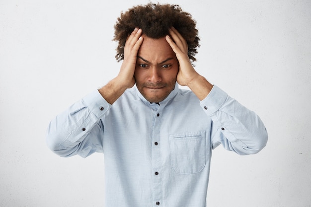 Jeune homme hipster à la peau sombre portant une chemise blanche tenant les mains sur la tête à désespérément vers le bas