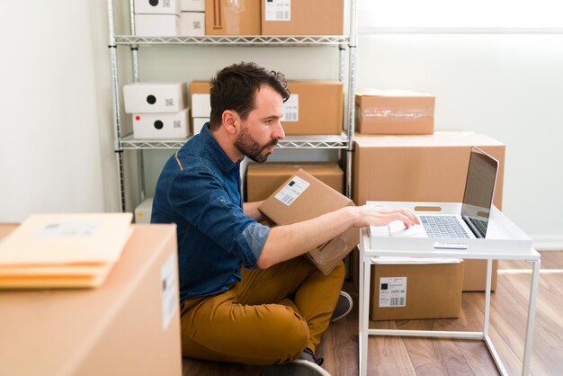 Jeune homme hispanique assis sur le sol de son bureau tout en tenant un paquet et en tapant sur l'ordinateur portable pour vérifier une commande client de la boutique en ligne