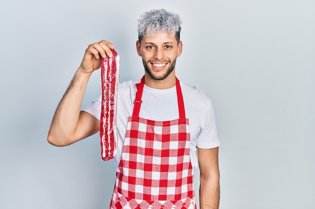 Photo gratuite jeune homme hispanique aux cheveux teints modernes portant un tablier tenant un steak de boeuf cru à la position positive et heureuse et souriant avec un sourire confiant montrant les dents