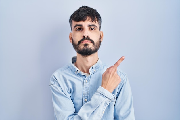 Jeune homme hispanique avec barbe debout sur fond bleu pointant avec le doigt de la main sur le côté montrant la publicité, le visage sérieux et calme