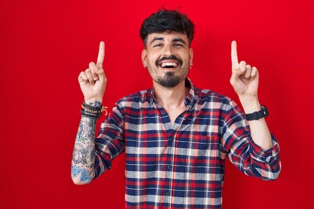 Jeune homme hispanique avec barbe debout sur fond rouge souriant étonné et surpris et pointant vers le haut avec les doigts et les bras levés.