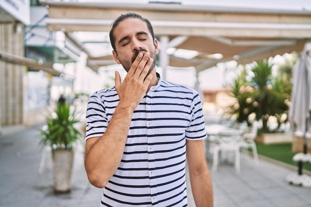 Photo gratuite jeune homme hispanique avec barbe à l'extérieur de la ville ennuyé bâillant fatigué couvrant la bouche avec la main. agitation et somnolence.